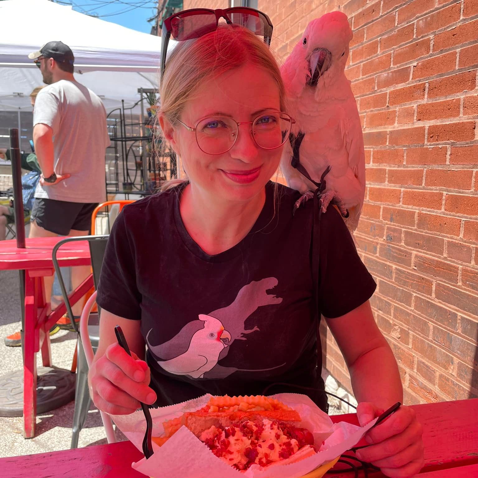 A woman wearing a T-shirt featuring a parrot sits at an outdoor table with a white cockatoo perched on her shoulder.